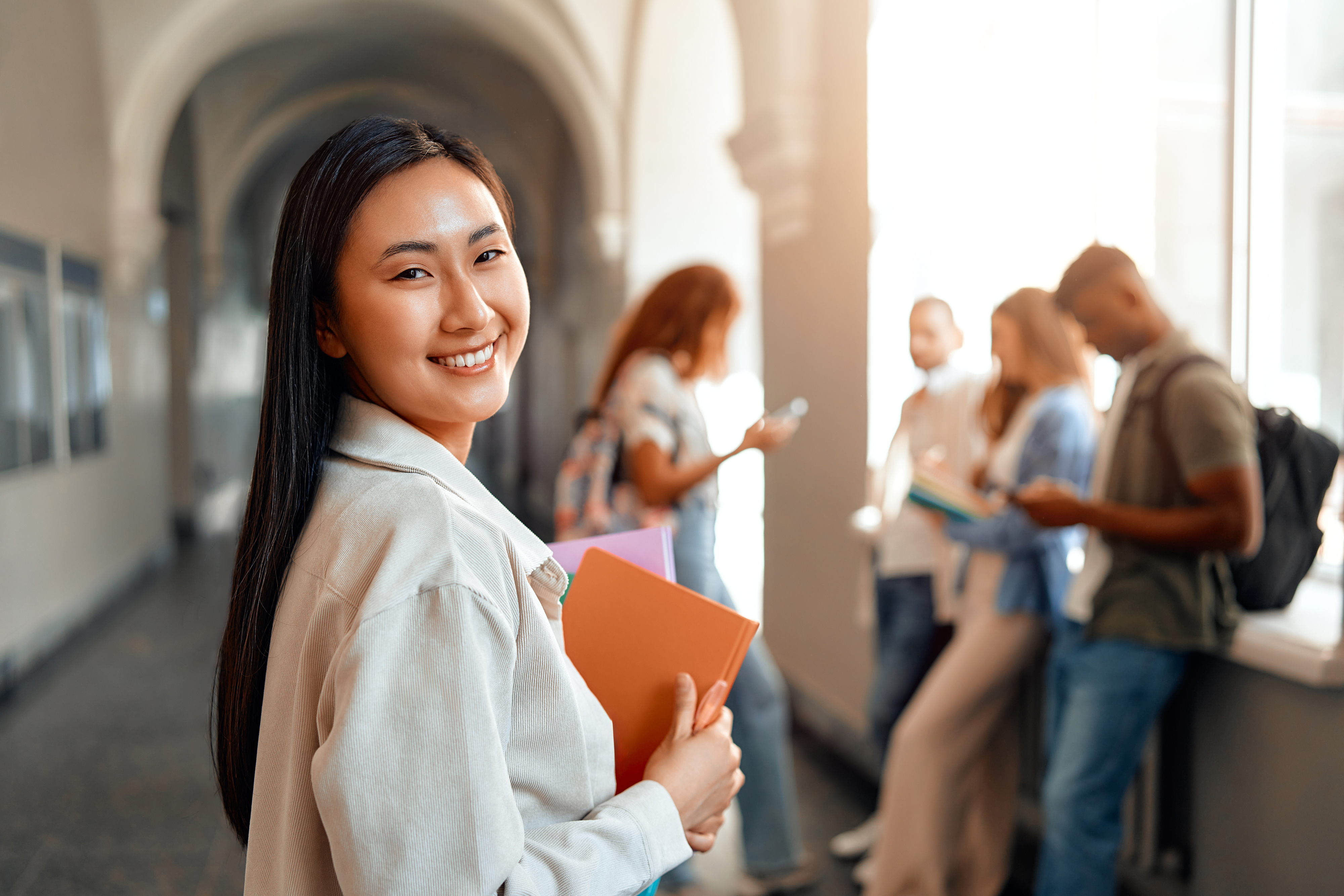 A student, holding notebooks, smiles at the camera in a hallway with several other students chatting and looking at their phones in the background
