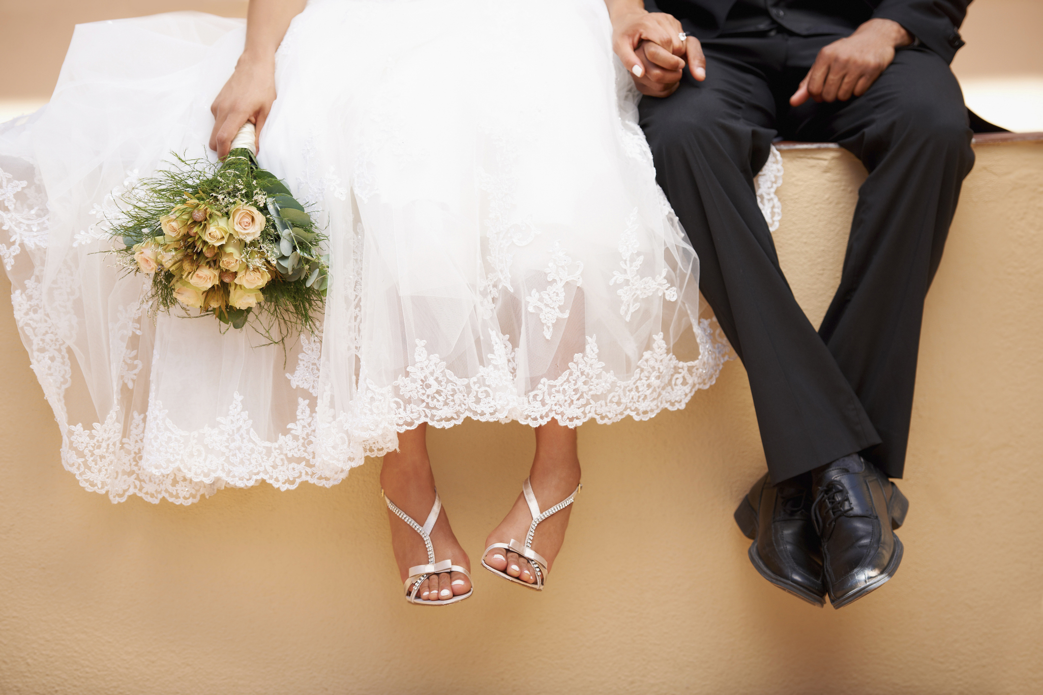 A bride and groom sit holding hands, with the bride's white lace dress and bouquet of yellow flowers visible, and the groom in a black suit and shoes