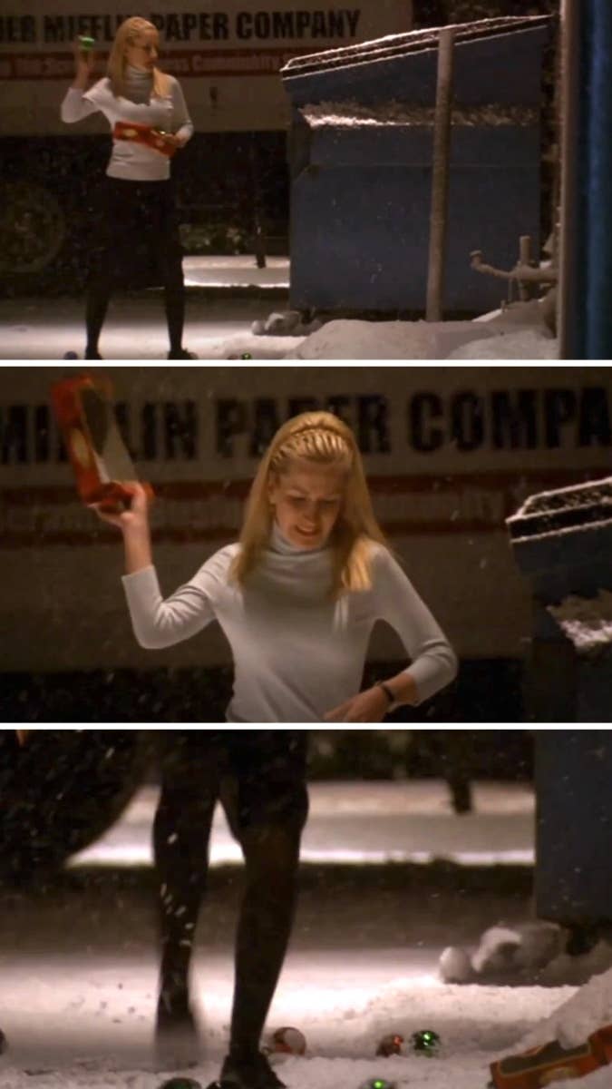 Reese Witherspoon, in a white top and black skirt, smashes Christmas ornaments outside near a truck in a snow-covered area