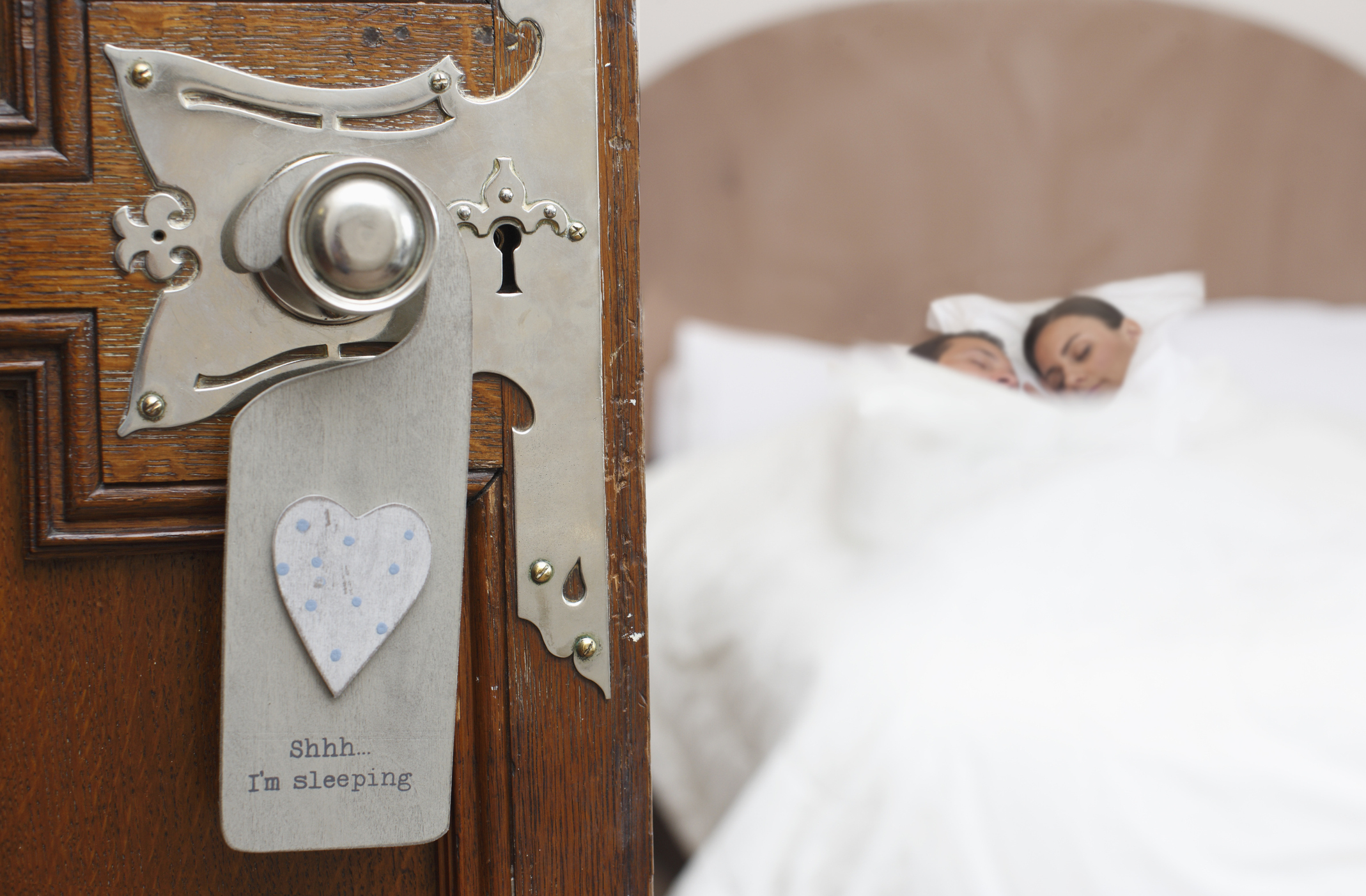 A couple is asleep in bed, visible through a partially open door. A &quot;Shhh&hellip; I'm sleeping&quot; sign hangs on the door handle