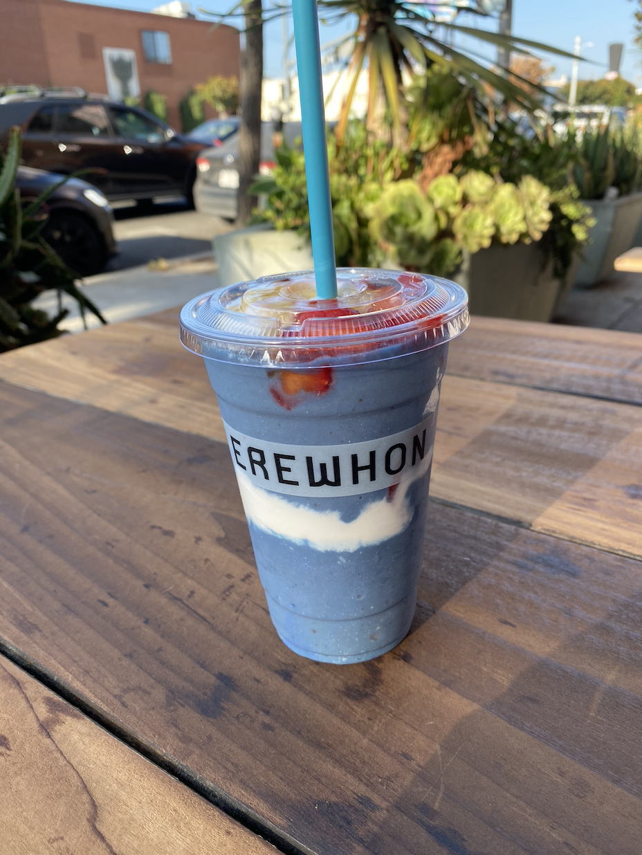 A purple layered smoothie in a plastic cup with a blue straw, labeled "EREWHON", placed on a wooden table outdoors near plants and parked cars