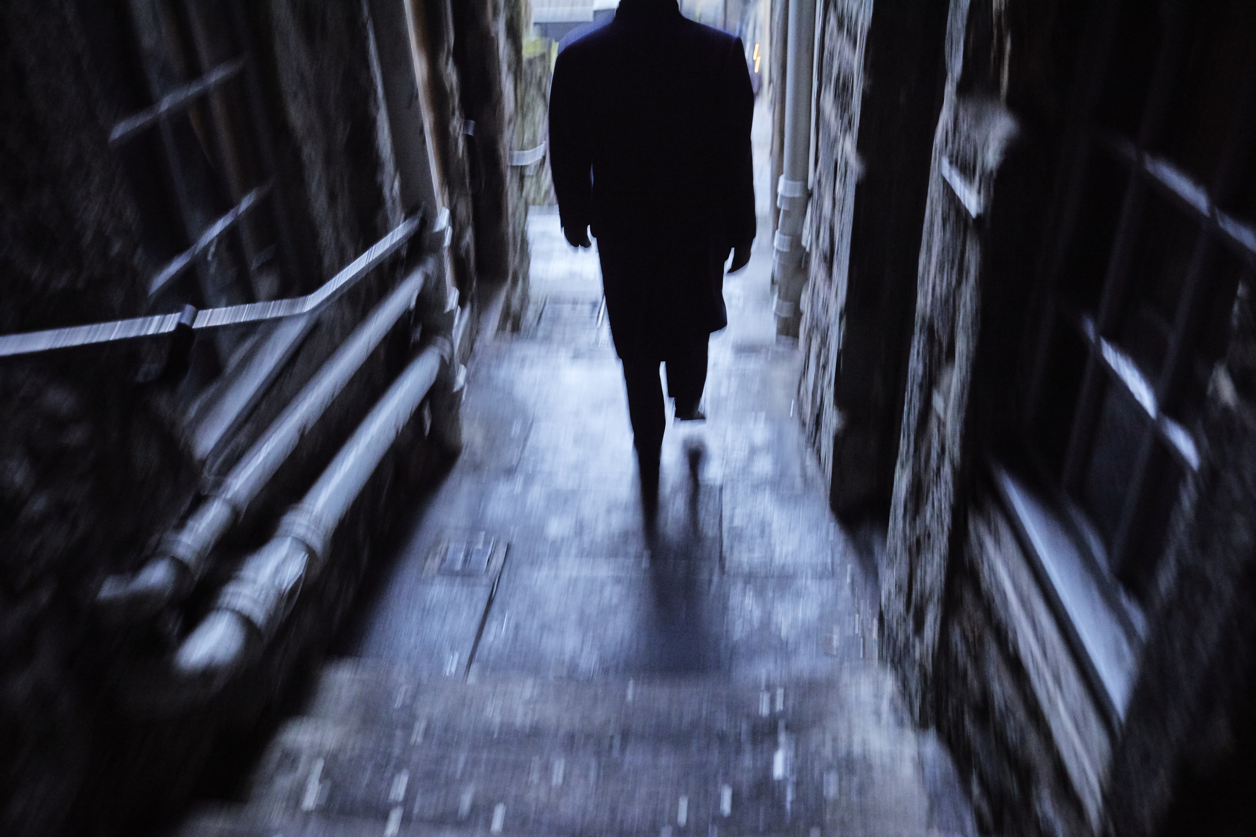 A silhouetted person in a coat walks down a narrow, dimly-lit stone alley