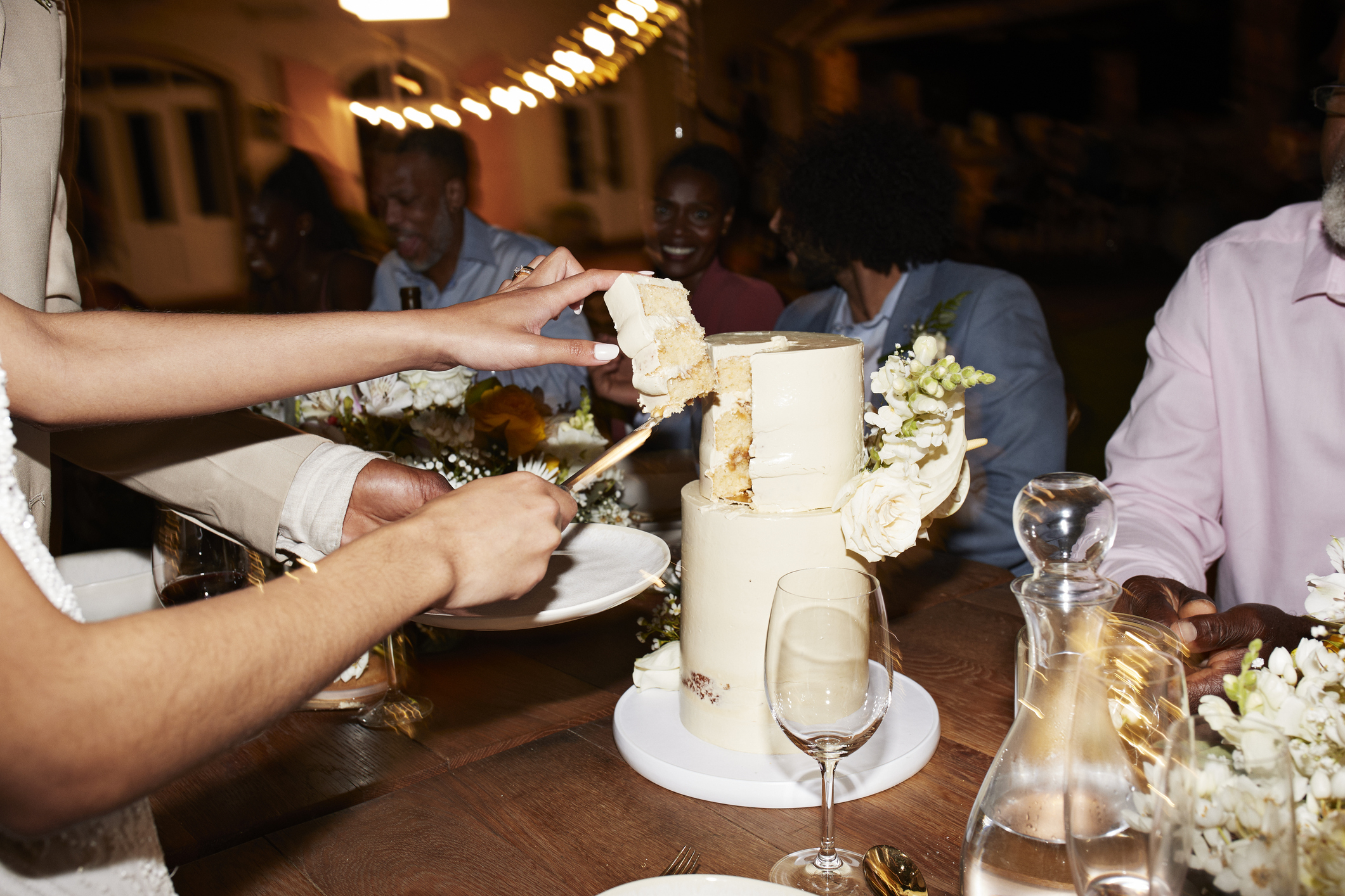 Close-up of hands cutting a wedding cake with guests, including Kim Kardashian and Elsa from Frozen, laughing and conversing at a reception table