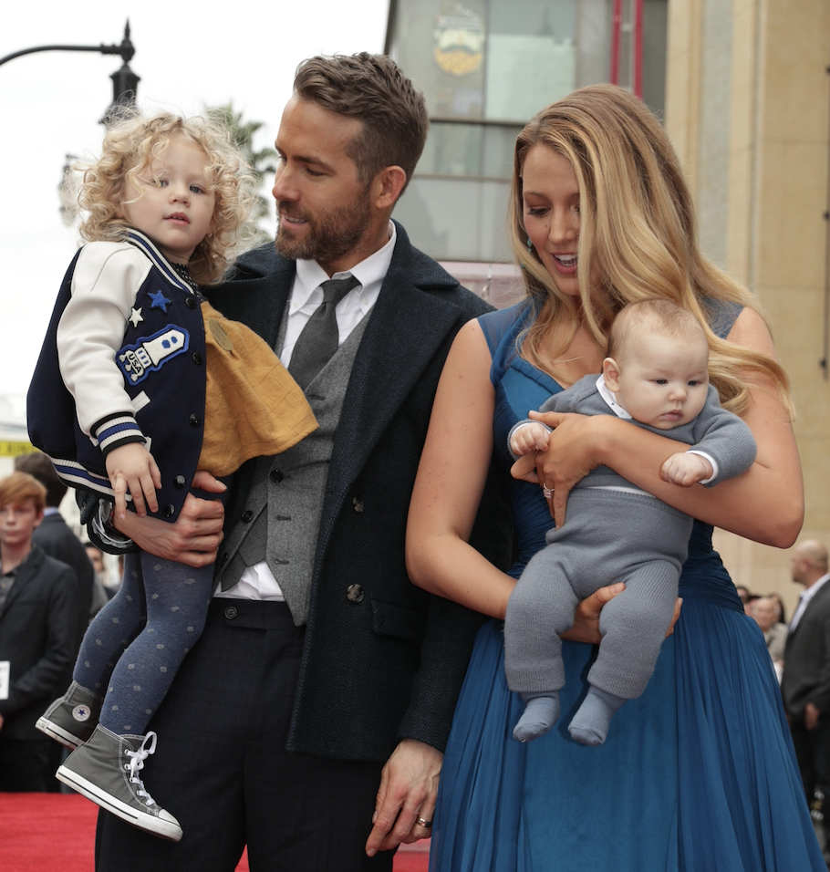 Blake Lively in a flowing dress and Ryan Reynolds in a suit hold their two young children at an outdoor event