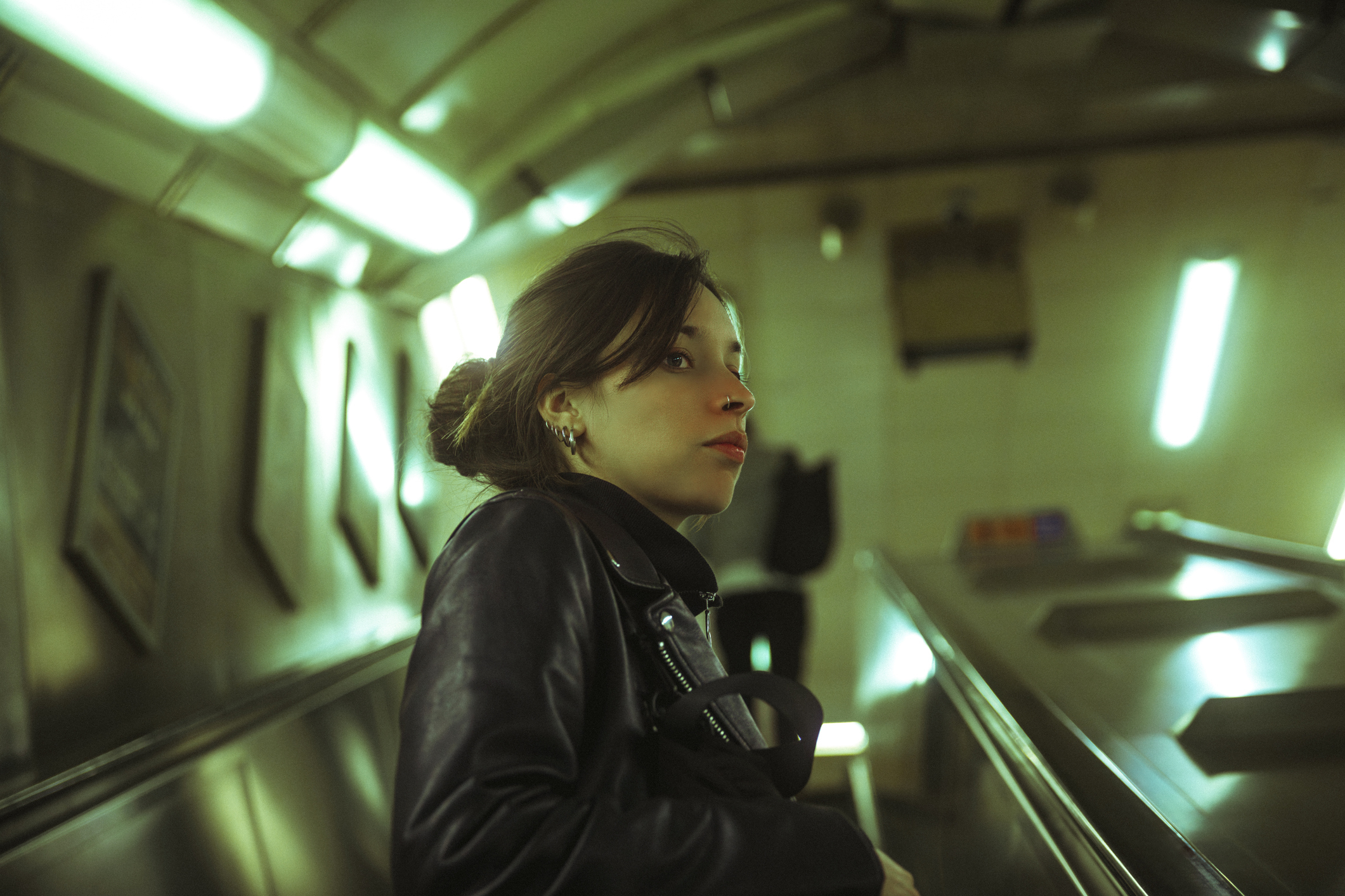 A woman in a subway station is standing on an escalator, looking to the side, wearing a leather jacket