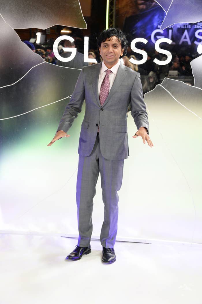 M. Night Shyamalan in a grey suit and pink striped tie, poses with arms slightly extended at the "Glass" movie premiere
