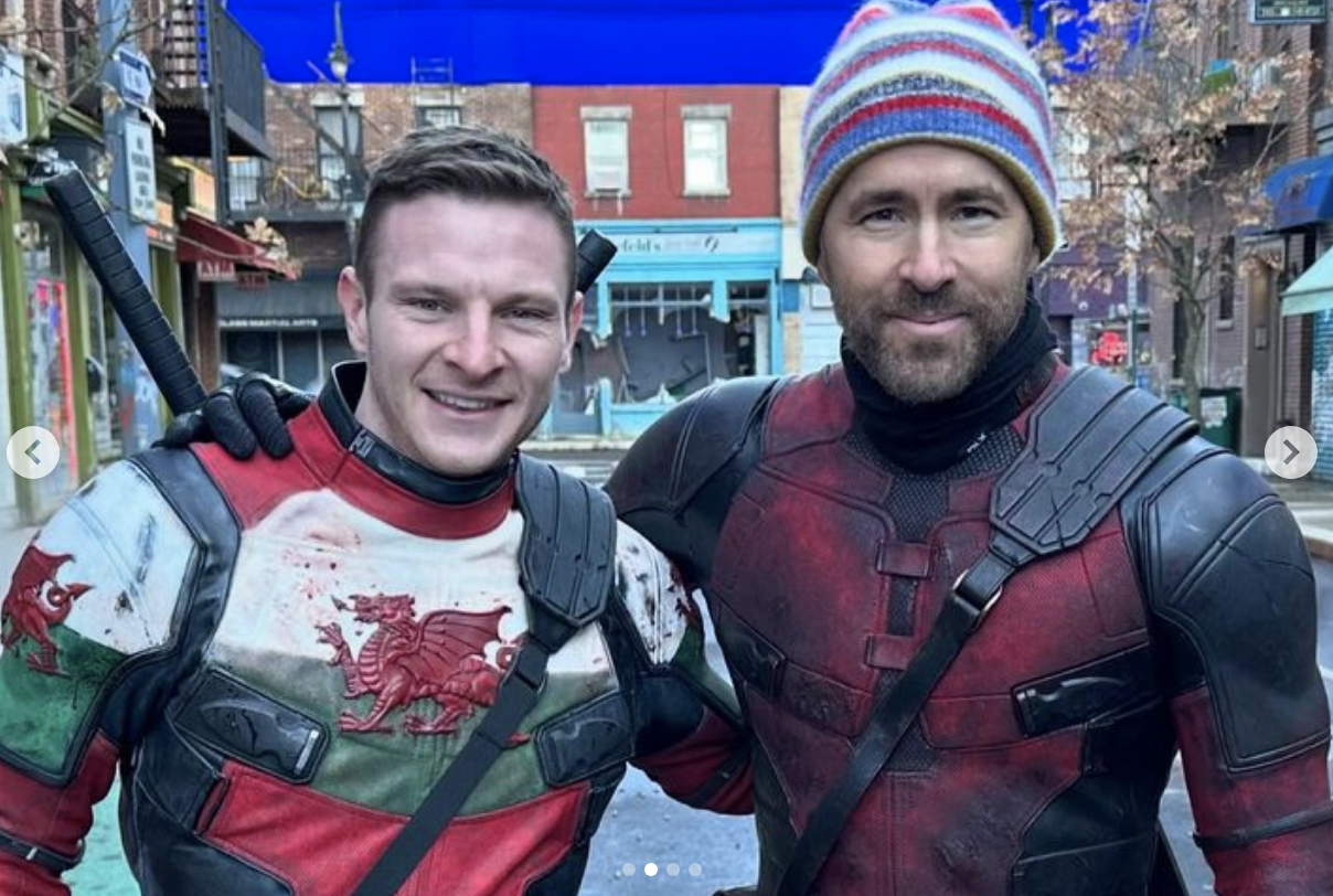 Ryan Reynolds in Deadpool costume posing with stunt double on an urban set. Both wearing protective gear and smiling at the camera