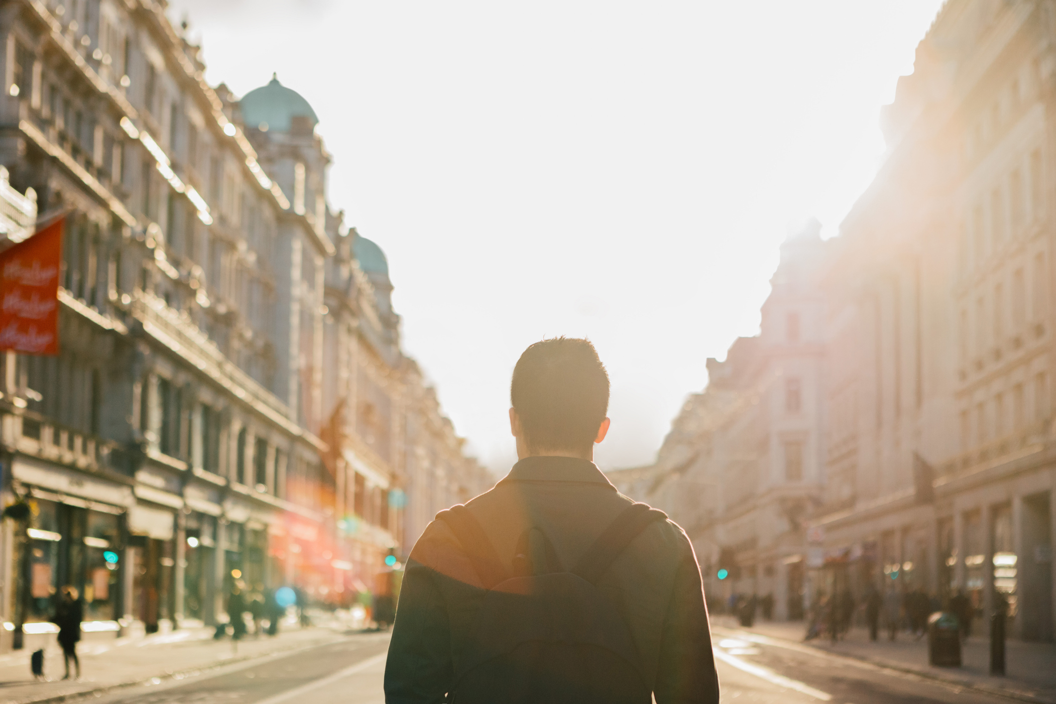 Person standing in the middle of a city street with tall buildings on either side, facing away from the camera. Sunlight bathes the scene