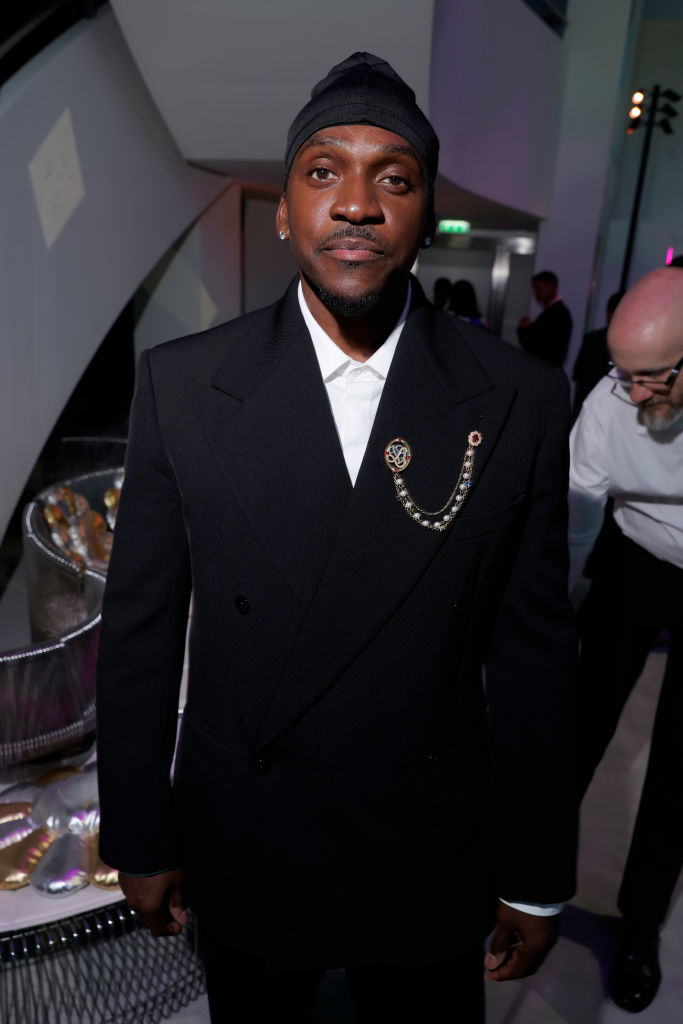 Man in a black suit with a decorative chain and black beanie, standing indoors. A man with glasses is in the background. Names unknown