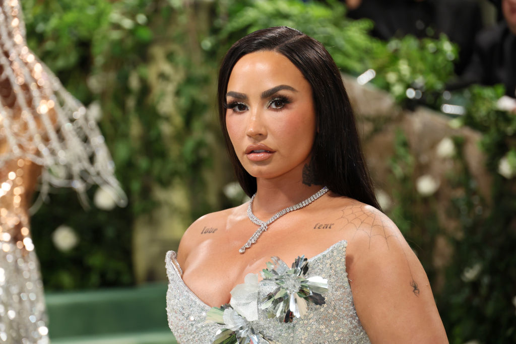Demi Lovato at a red carpet event wearing an elegant, sparkling dress with floral embellishments and a diamond necklace