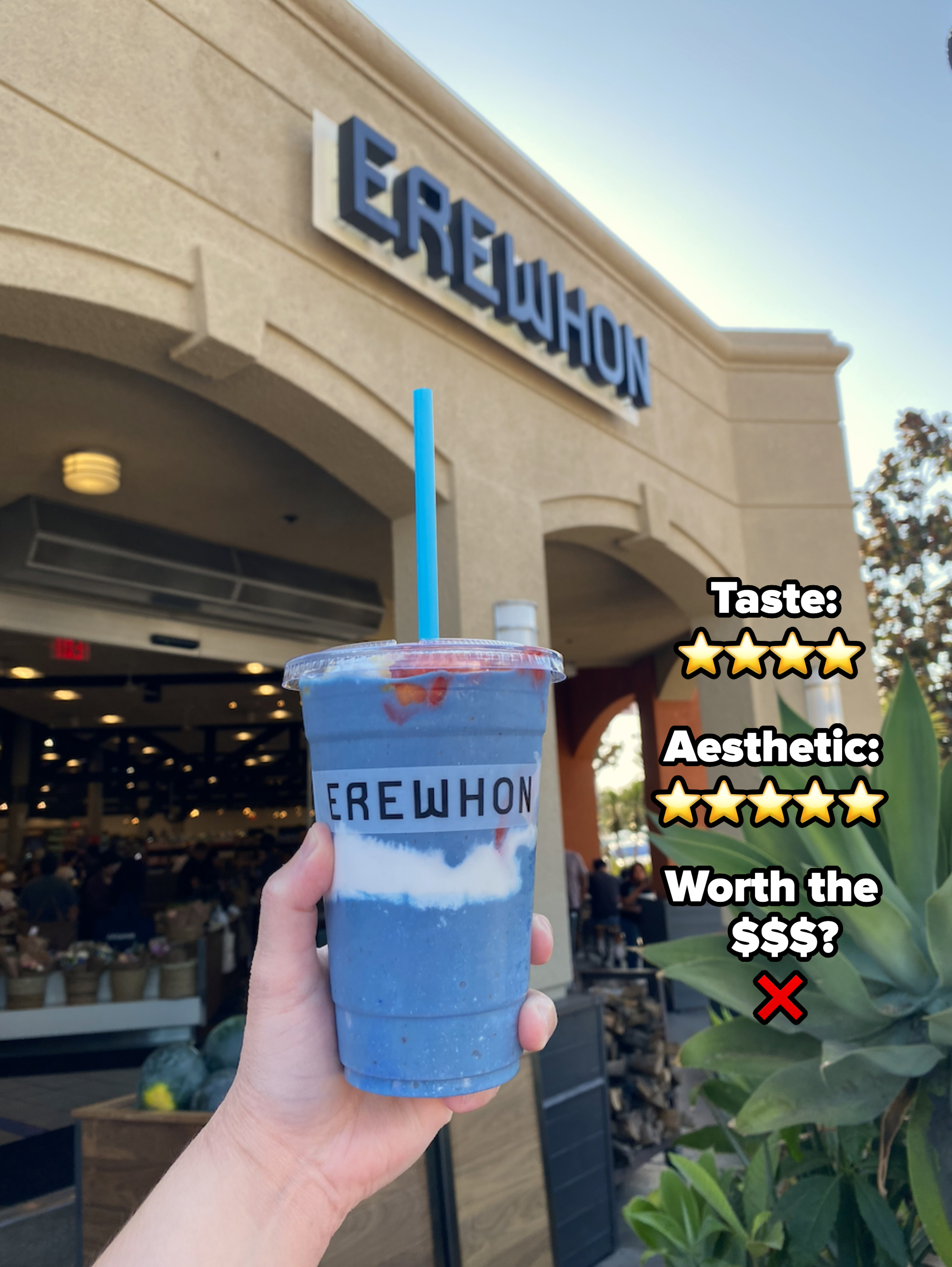 A hand holds a smoothie with a blue straw in front of Erewhon market