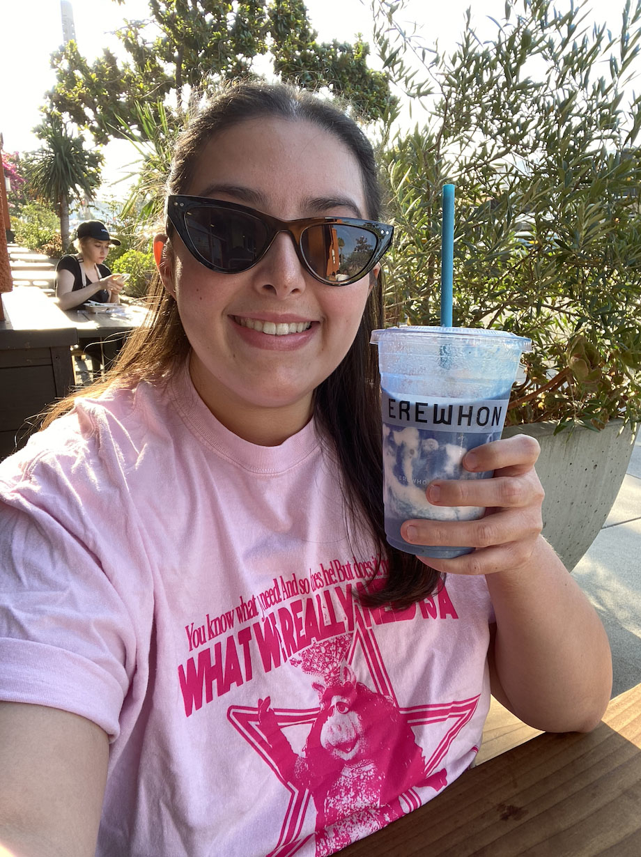 Person smiling at a cafe, wearing sunglasses and a pink "What Would Alyssa Milano Do?" shirt, holding a drink from Erewhon. Other patrons are in the background