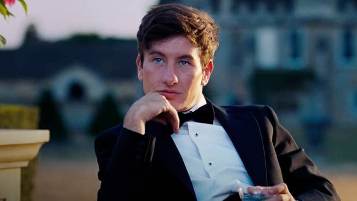 Barry Keoghan is sitting outdoors in a black tuxedo and white dress shirt, holding a glass, and gazing intently into the distance