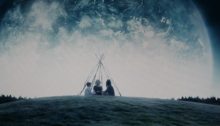 Three people sit in front of a simple wooden tipi structure on a grassy hill, with a large Earth-like planet visible in the sky above them