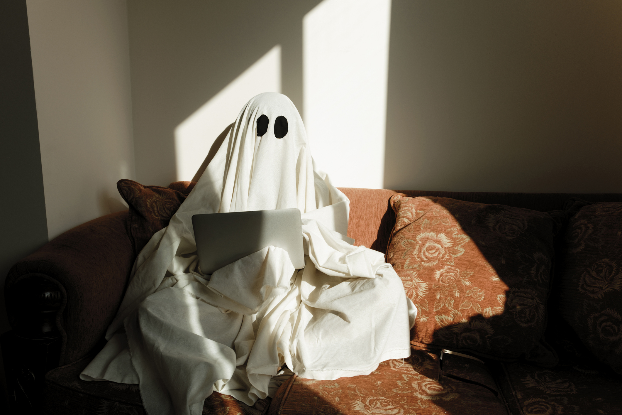Person dressed as a ghost, covered with a sheet with black eye holes, sitting on a sofa using a laptop in a brightly lit room