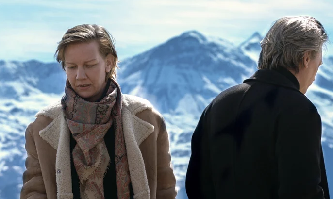 Sandra Hüller and Bruno Ganz in a snowy mountain scene, dressed warmly with Sandra Hüller wearing a scarf and coat while Bruno Ganz faces away in a dark coat