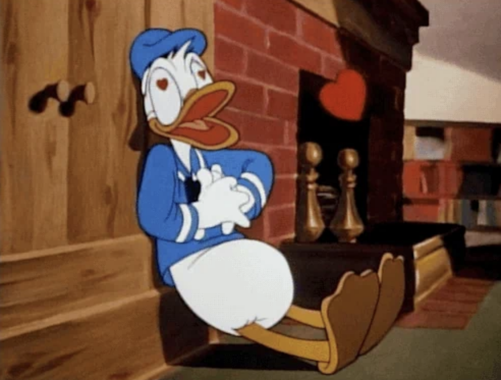 Donald Duck, dressed as a sailor, leans against a brick wall with heart eyes and a red heart above his head, indicating he's in love