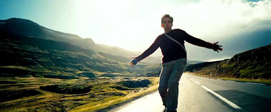 Ben Stiller rides a skateboard along a scenic, open road with mountains in the background. He appears to be enjoying the ride, arms stretched out
