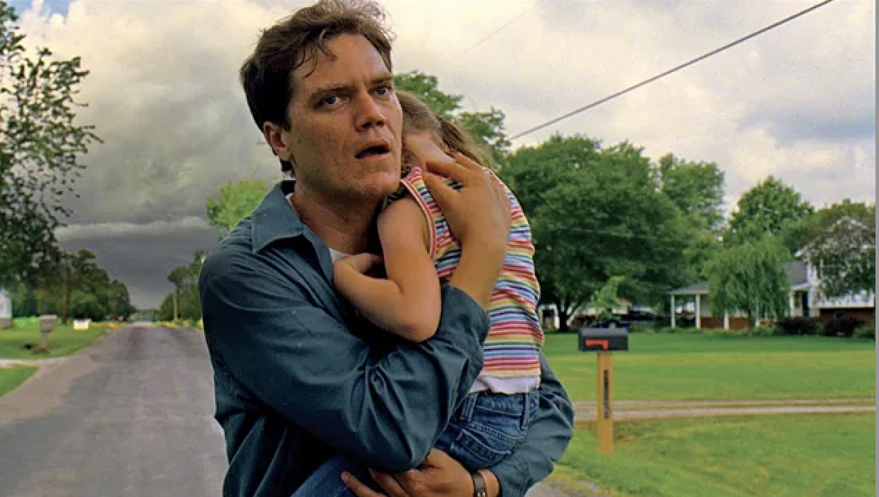 Michael Shannon holds a young girl in his arms, looking concerned, in a suburban street with trees and houses in the background