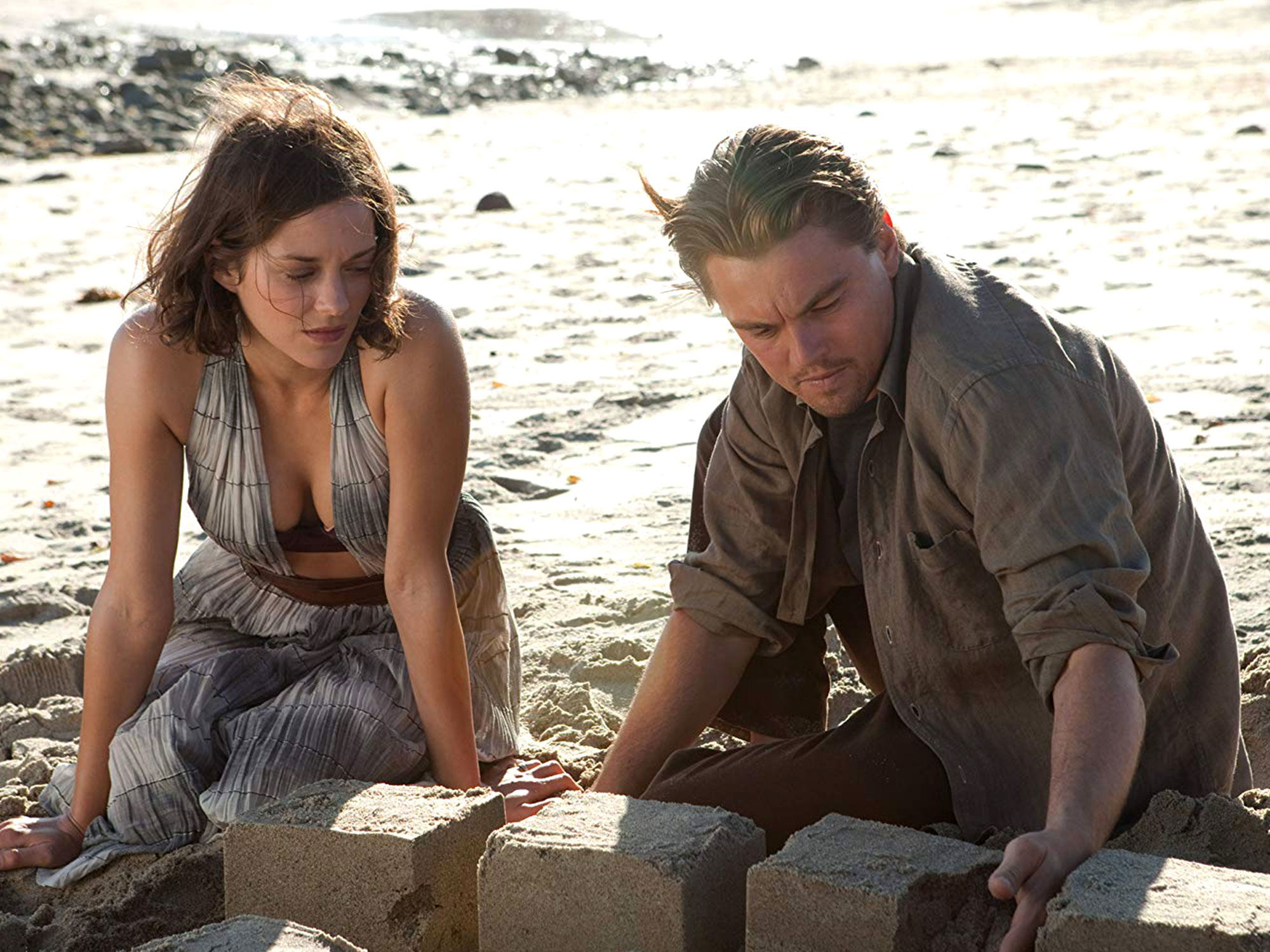 Marion Cotillard and Leonardo DiCaprio are sitting on a beach, focusing on constructing a sand structure with serious expressions