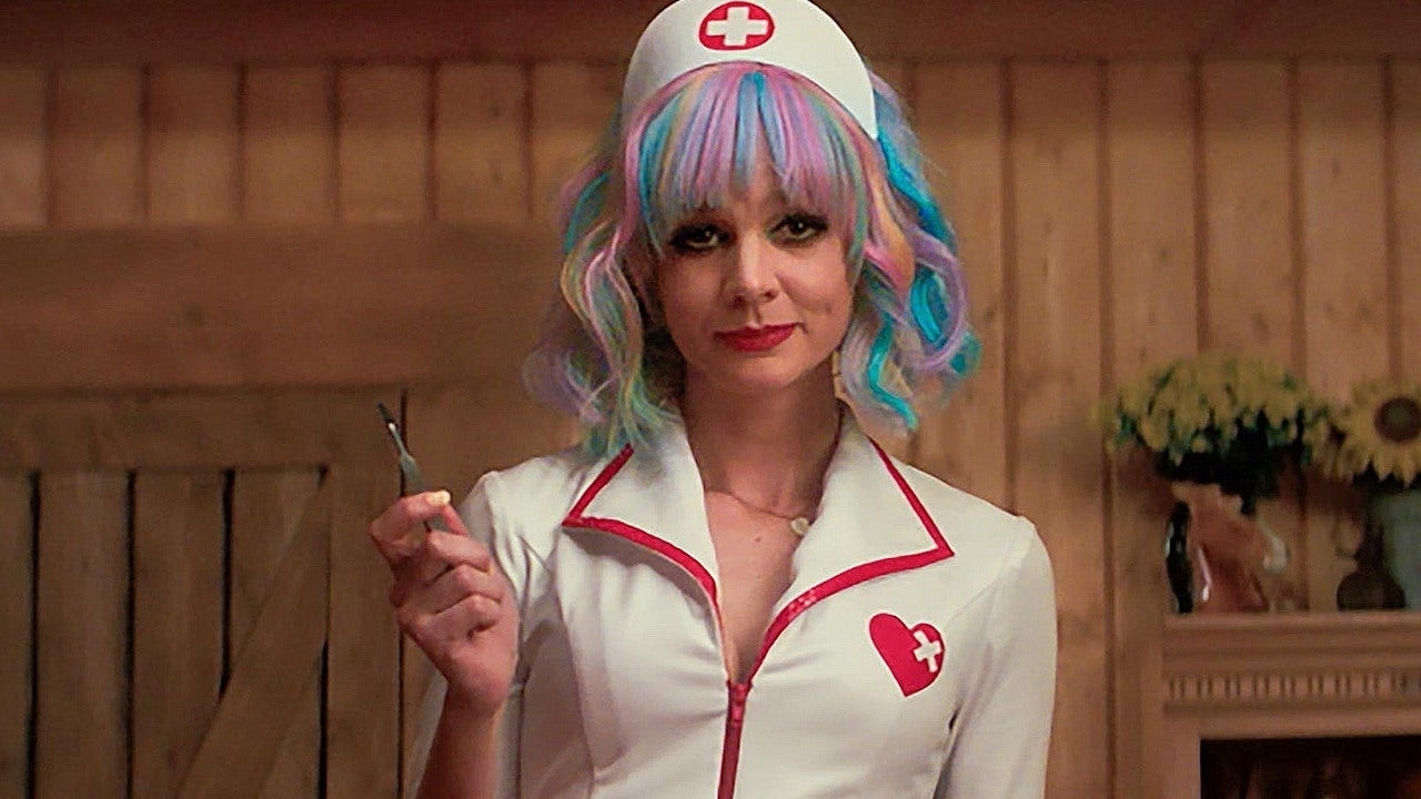 Carey Mulligan in a nurse costume with a syringe, featuring a white outfit with red trim and a heart with a cross emblem. She is in a wooden setting