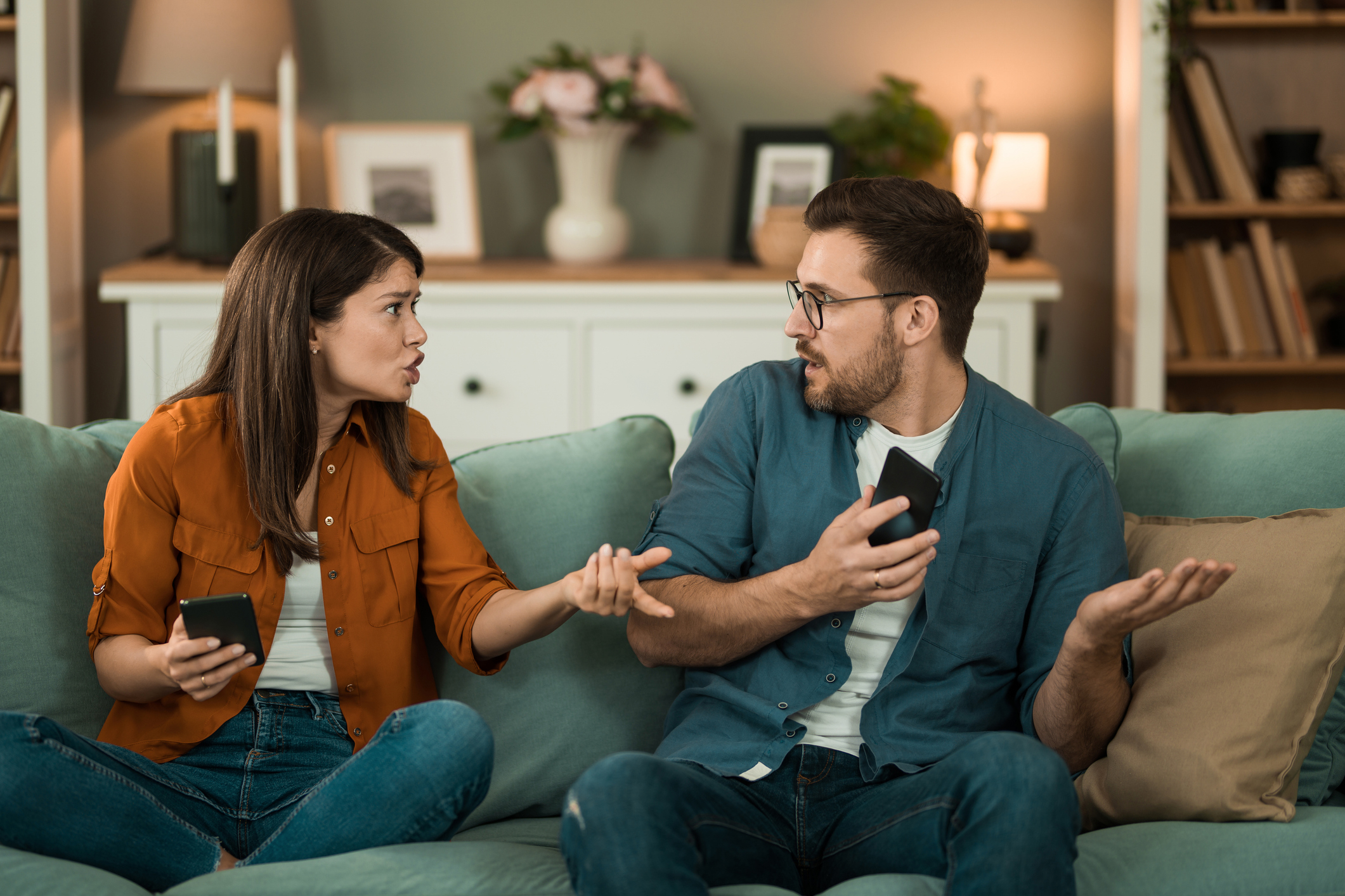 A woman and a man sit on a couch arguing while holding smartphones. There is a blurred background of shelves and a cabinet with decor items