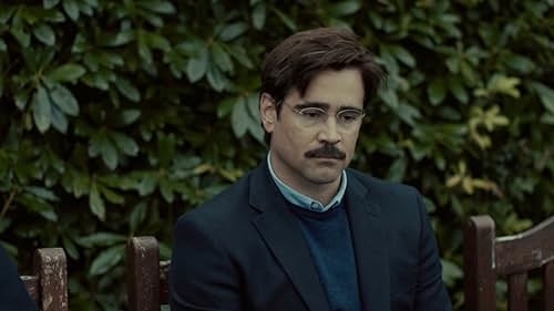 Colin Farrell, wearing glasses and a suit, sits on a wooden bench against a leafy background, looking contemplative