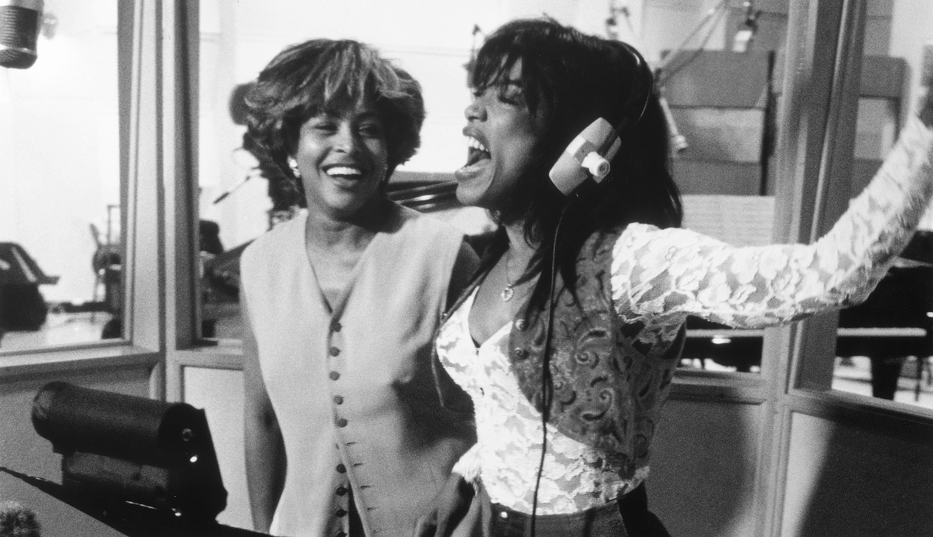 Tina Turner and Angela Bassett in a recording studio, smiling and laughing; Turner wears a button-up vest, and Bassett wears a lace-sleeved top with headphones