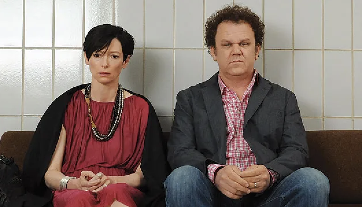 Tilda Swinton in a sleeveless dress with a necklace and John C. Reilly in a checkered shirt and blazer, sitting against a tiled wall
