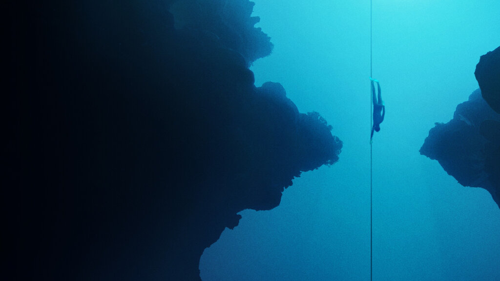 A free diver swims down a rope into the deep blue ocean, surrounded by large underwater rock formations