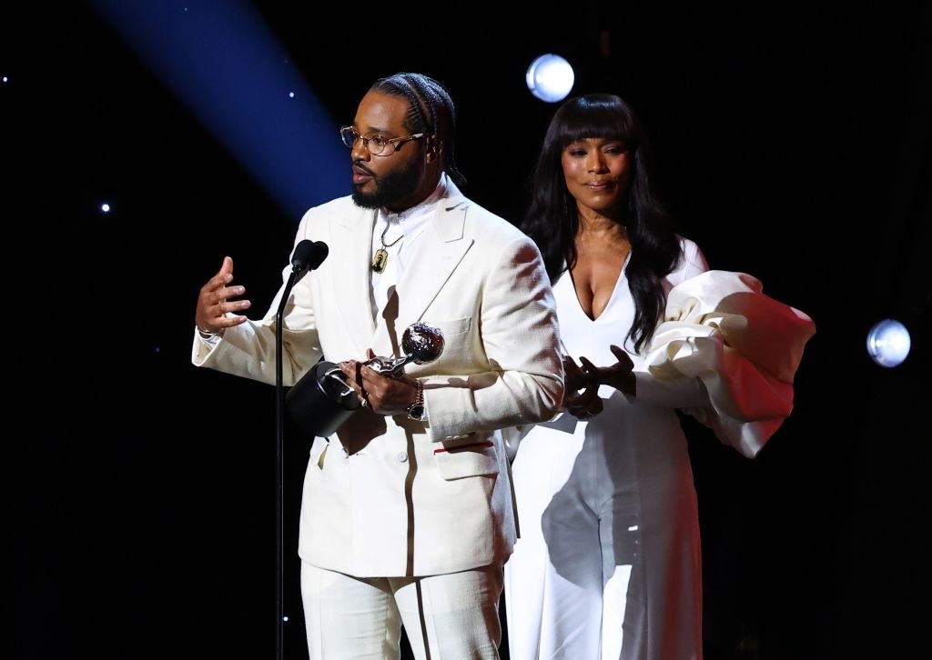 Ryan Coogler, dressed in a white suit, speaks at a podium while Angela Bassett, in a white dress with voluminous sleeves, stands behind him holding an award