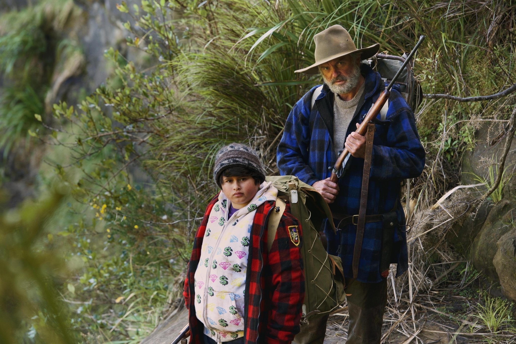 Julian Dennison and Sam Neill in outdoor gear, holding packs in a wilderness setting with dense vegetation