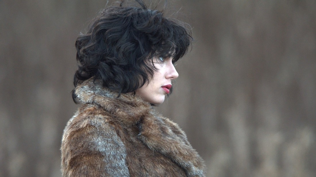 A woman with wavy black hair, wearing a fur coat, gazing contemplatively to the side against a blurred background