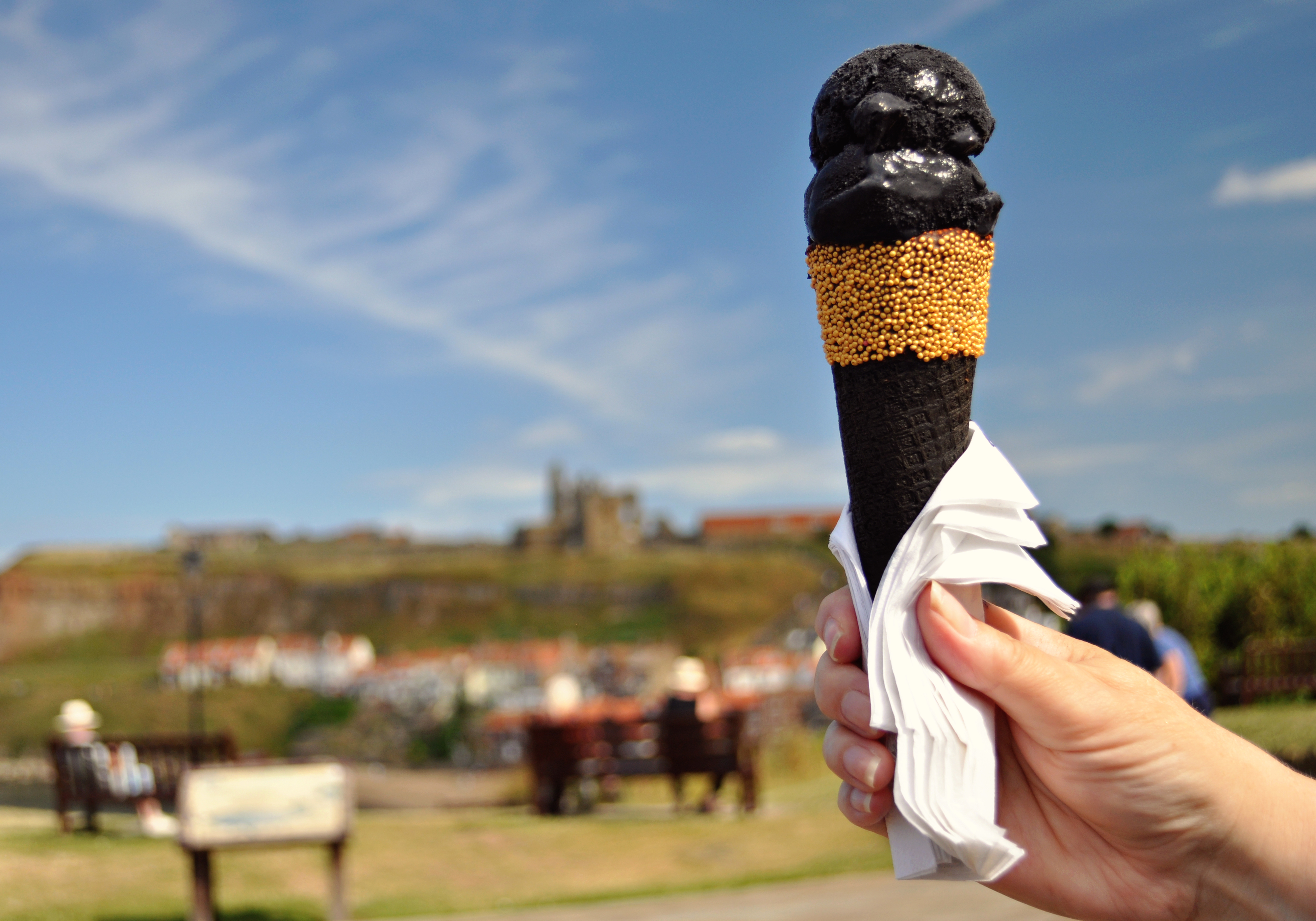A person holding a black ice cream cone with gold sprinkles in a park setting. People are sitting on benches in the background with a castle-like structure visible in the distance