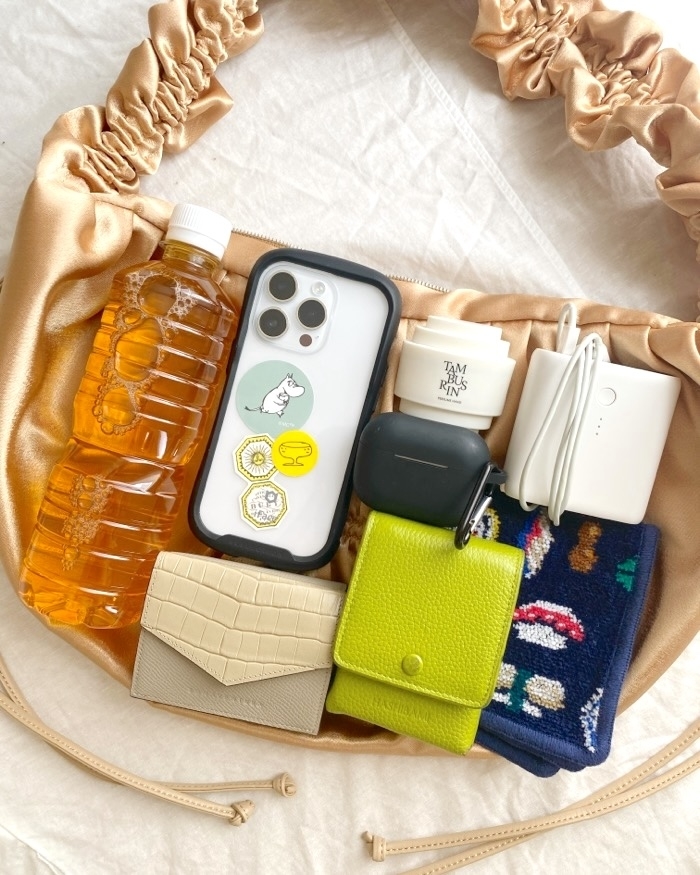 A satin handbag contains a bottle of tea, a smartphone with a cute animal sticker, a charging cable, an AirPods case, hand cream, a beige wallet, a lime green wallet, and printed socks