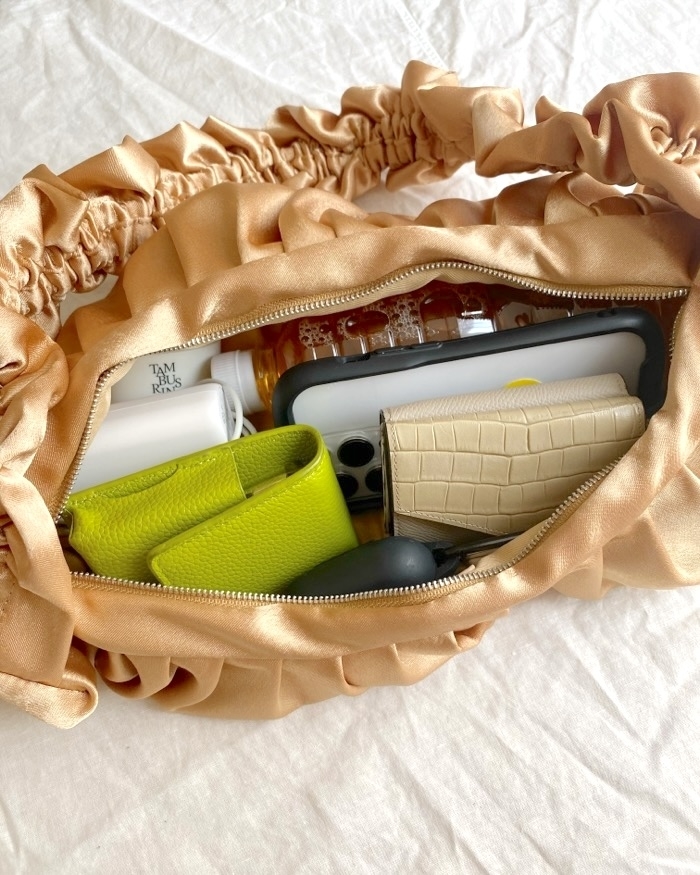 A satin handbag is open, revealing various items inside, including a green wallet, tan wallet, phone, face moisturizer, and a small bottle of liquid