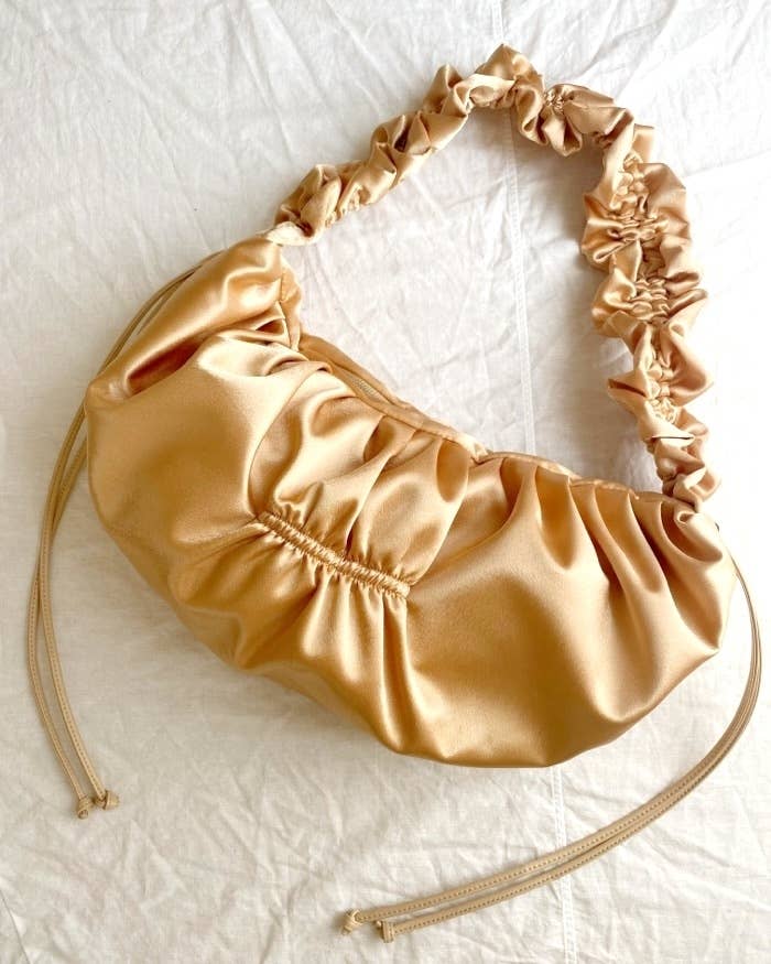 A satin ruched shoulder bag with drawstring accents, sitting on a white fabric surface. No text or people are present in the image