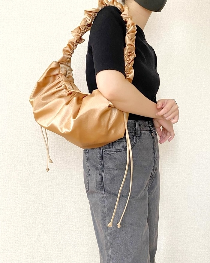 A person wearing a black top and grey jeans is holding a shiny, ruched handbag with drawstrings over their shoulder