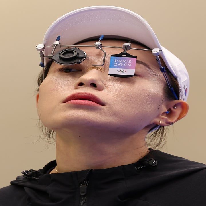 A person wearing a white cap and specialized shooting glasses with a tag reading "Paris 2024 OQ" on the right lens, part of an athletic event