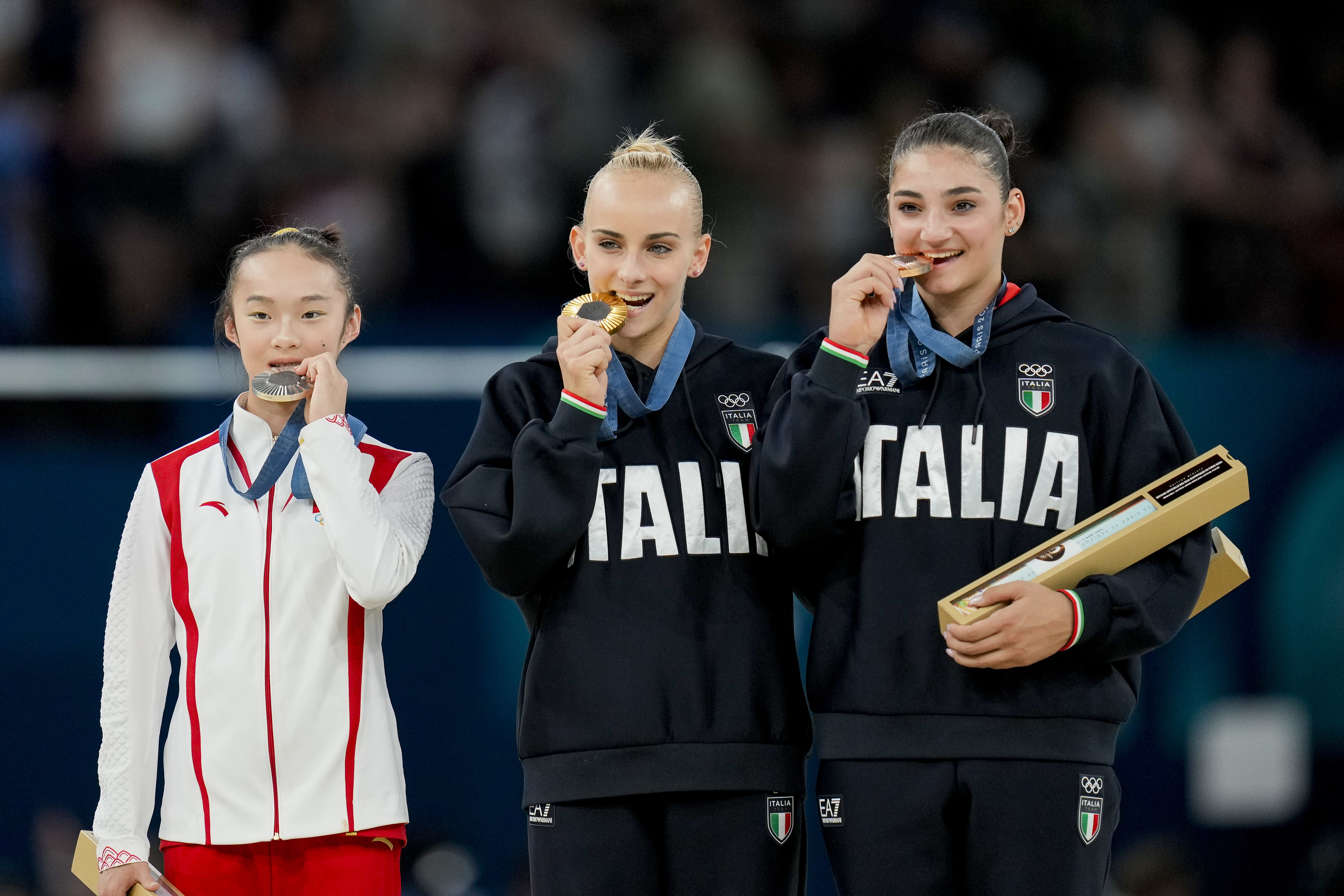 Three athletes, one in a white and red outfit, and two in Italian tracksuits with &quot;ITALIA&quot; text, pose with gold medals in their mouths