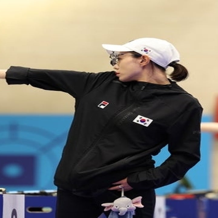 Shooter Jin Seok-hyeon in a black athletic jacket and white cap aims a pistol during a competition. Another person seen in the background adjusting equipment