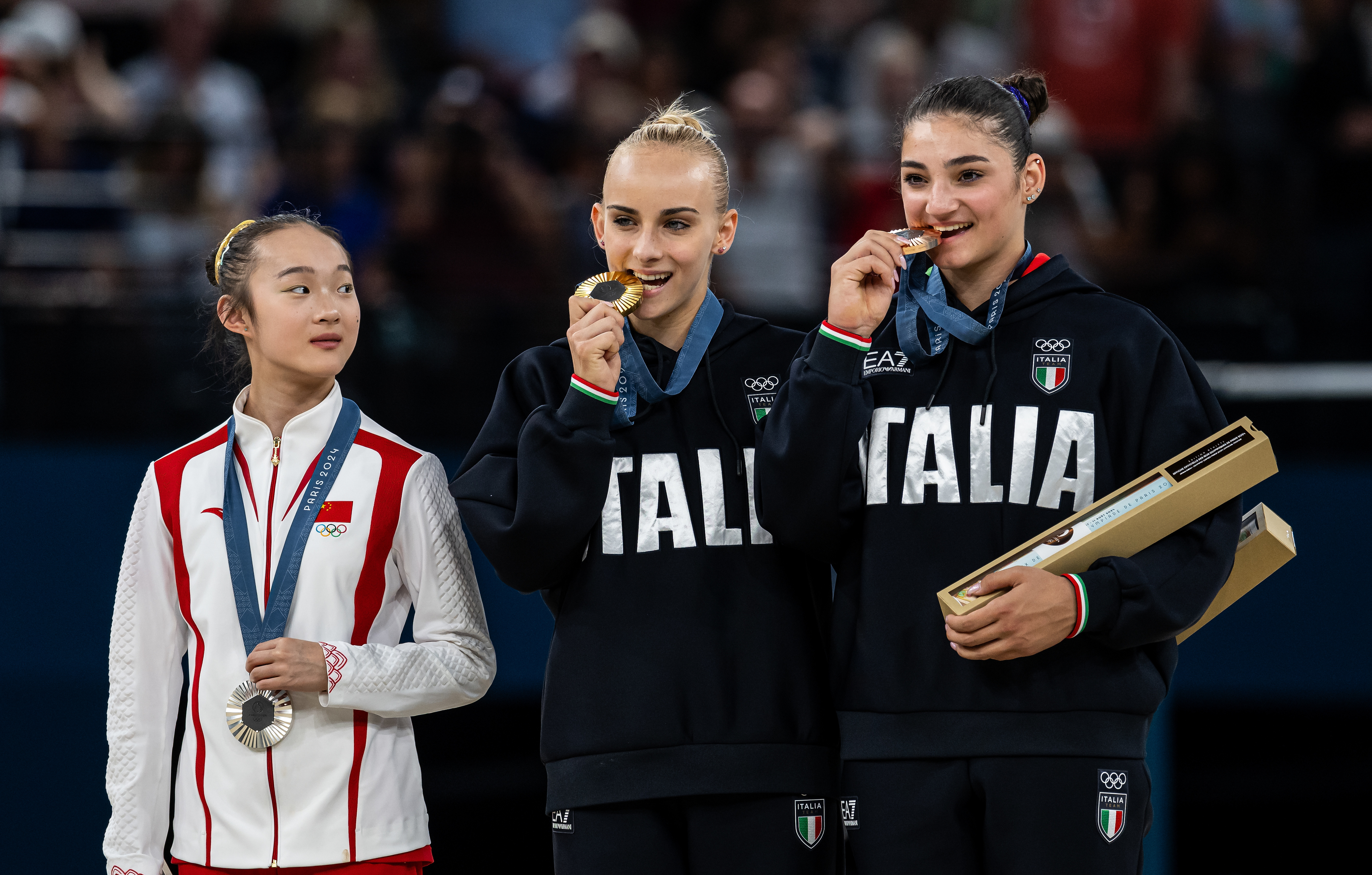 Olympic medalists pose on the podium: one athlete with a silver medal and two athletes wearing Italian tracksuits with gold medals