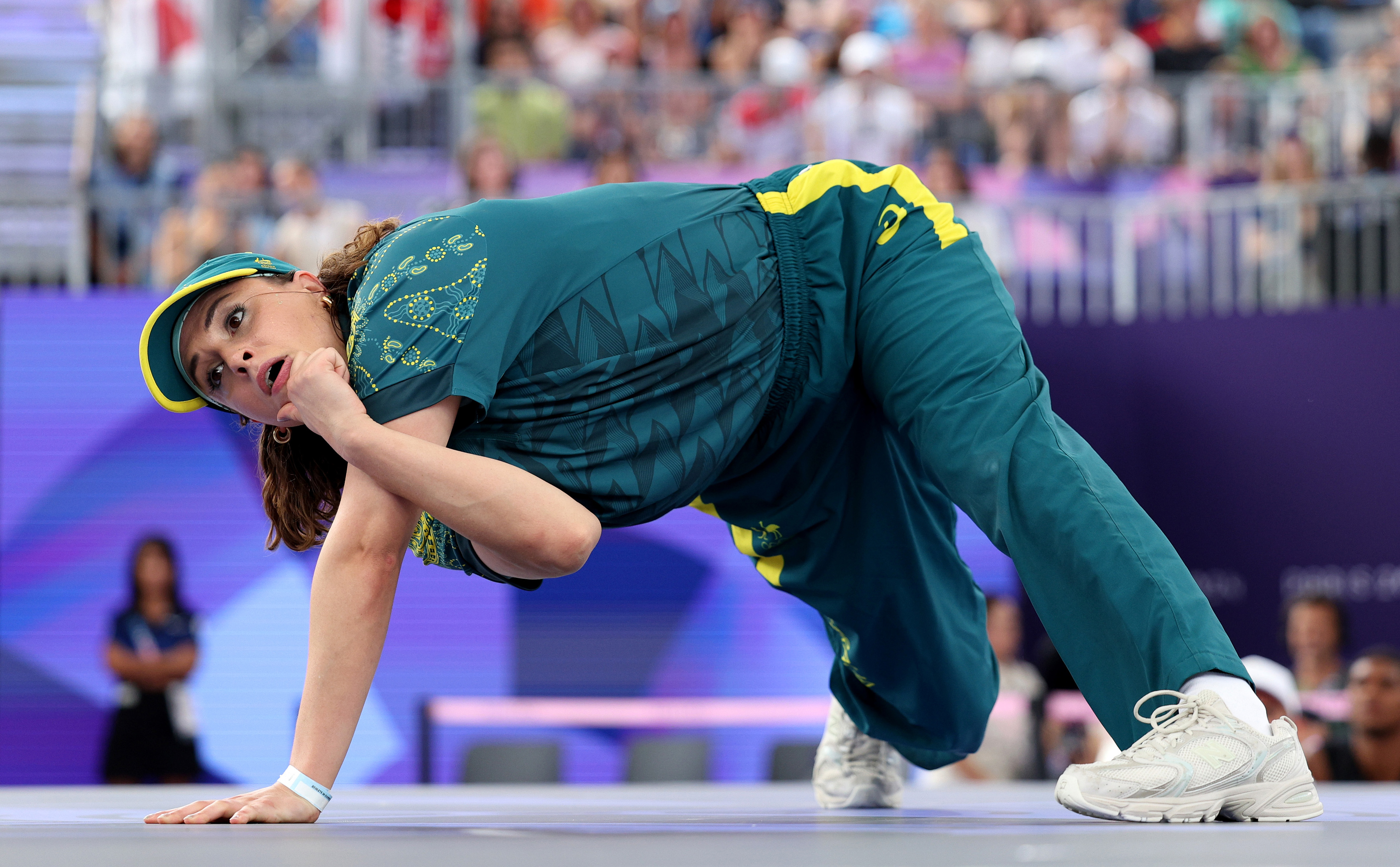 A person wearing a patterned shirt, cap, and sports pants is performing an intense dance move, bent down with one hand on the floor and the other at their chin