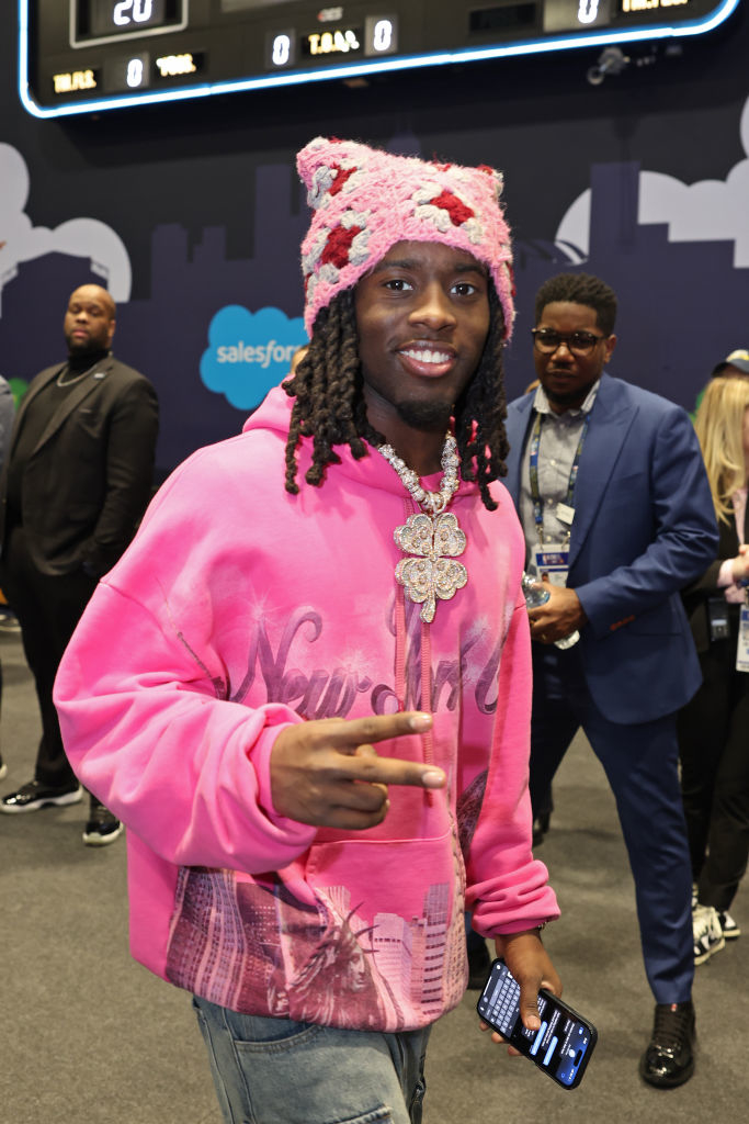 Kai Cenat flashes a peace sign while wearing a pink knit hat and a pink New York hoodie at a public event