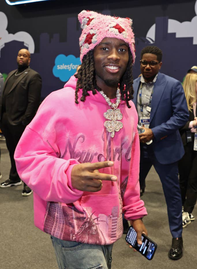 Kai Cenat flashes a peace sign while wearing a pink knit hat and a pink New York hoodie at a public event