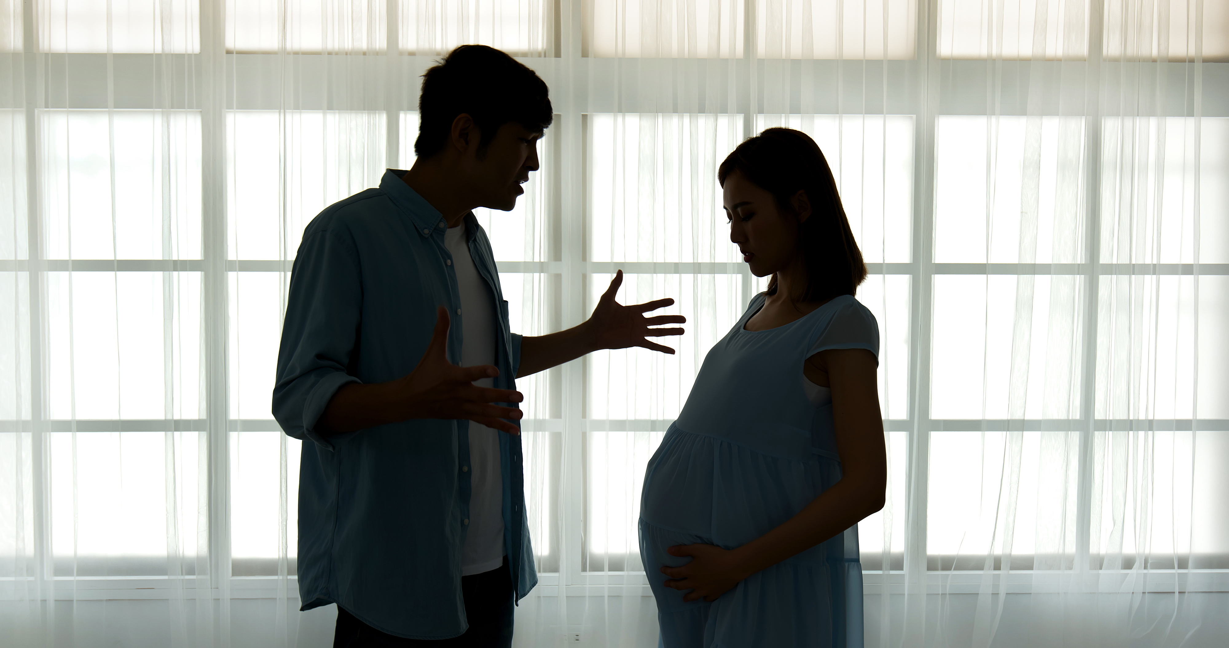 A pregnant woman and a man stand in front of a backlit window, facing each other. The man appears to be gesturing as he speaks to her