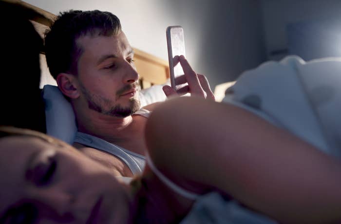 Man in bed looking at phone while woman sleeps beside him