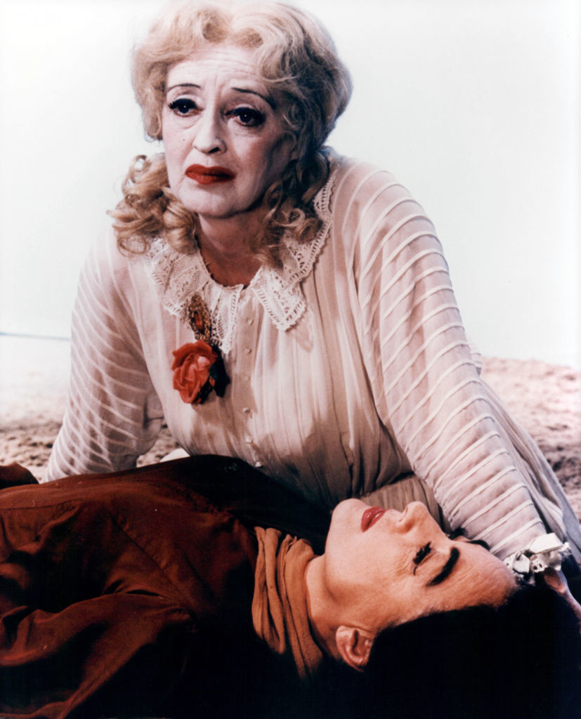 Bette Davis in a white, lace-collared blouse holds Joan Crawford, lying in her lap, in a dramatic scene from a film