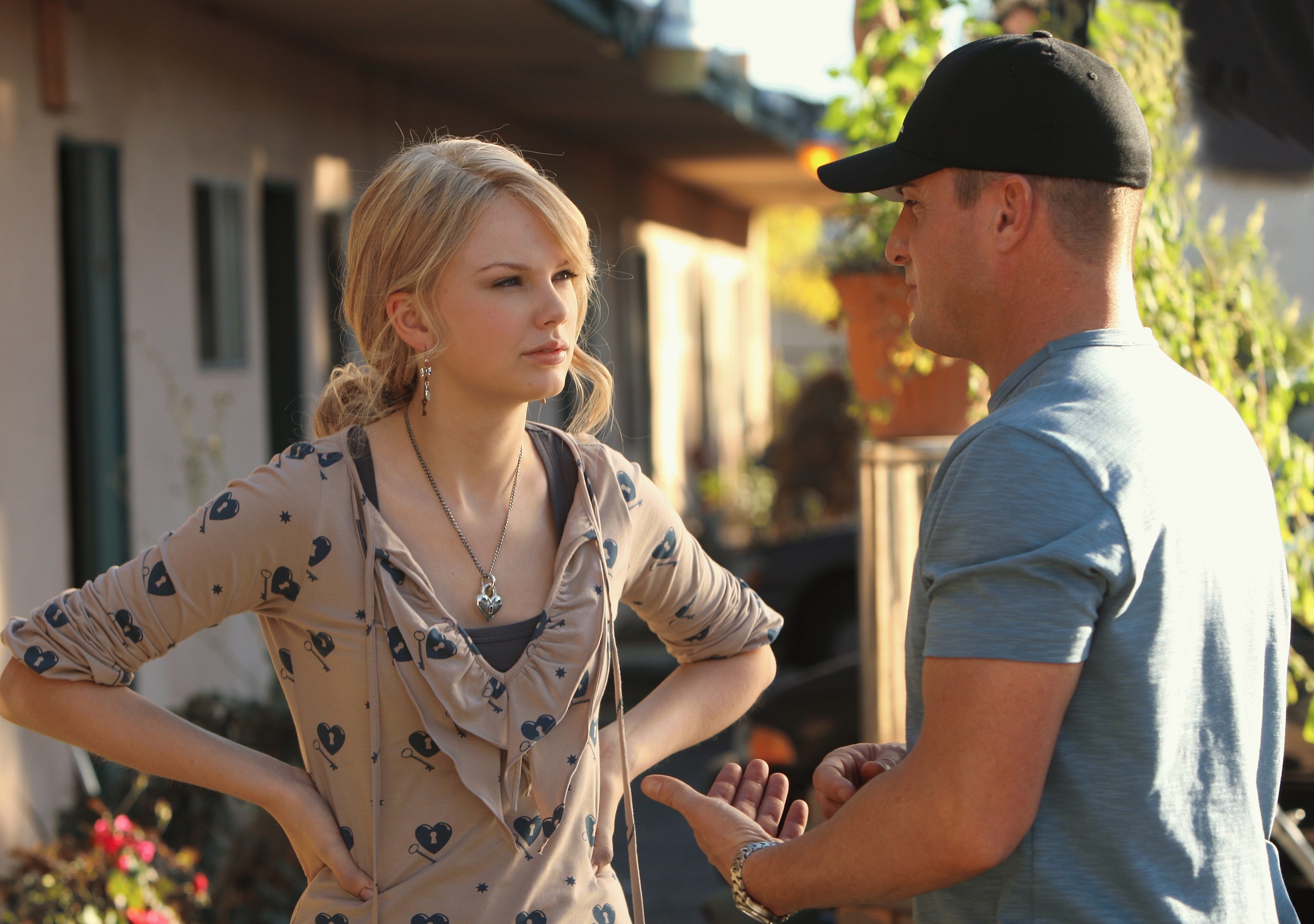 Taylor Swift in a patterned blouse with hands on hips, talking to a man in a cap and blue shirt