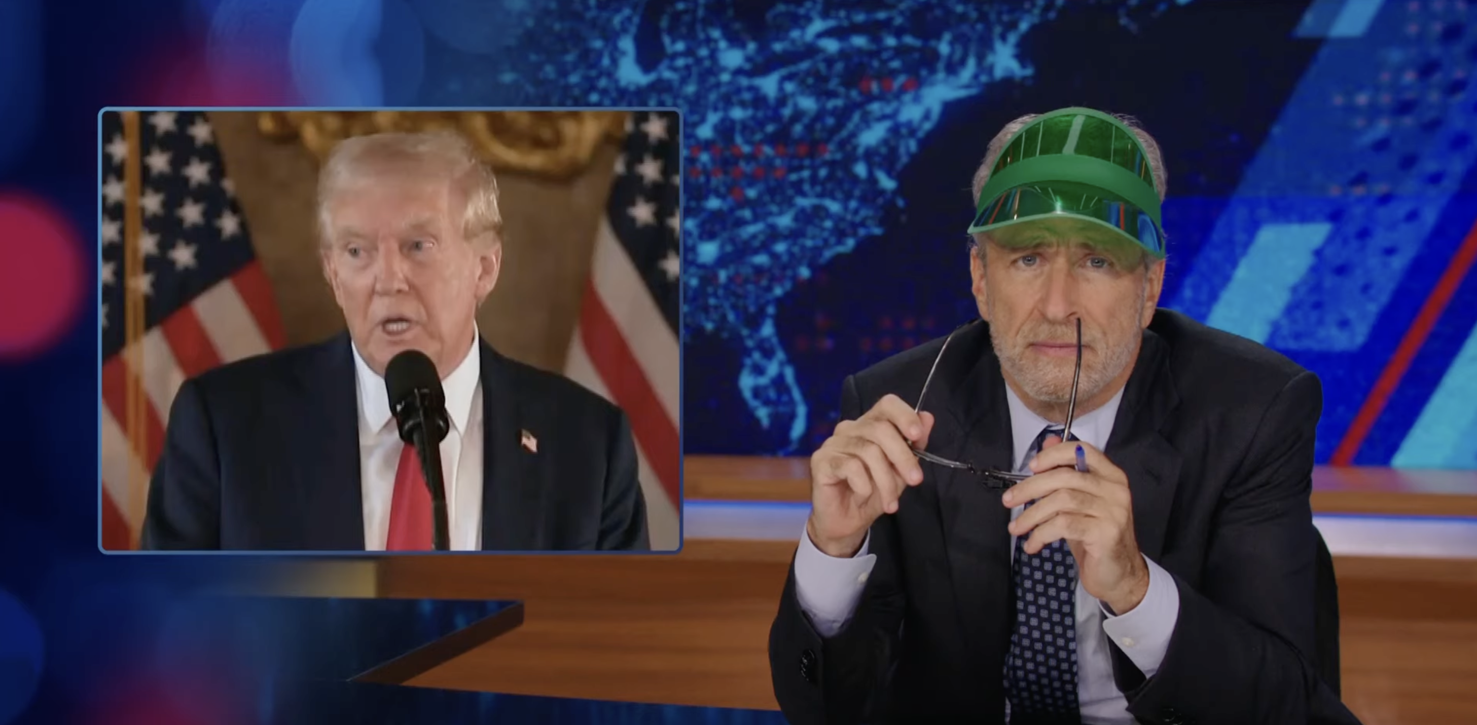 Donald Trump speaking on a screen while a man wearing a visor suit jacket and tie sits at a desk holding glasses